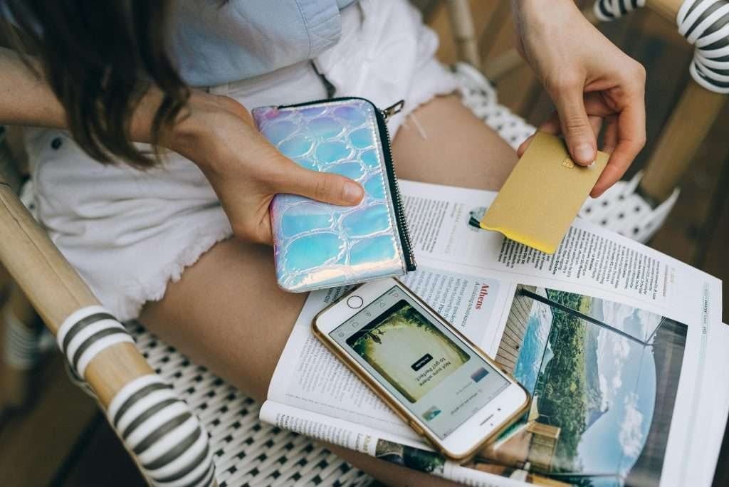 Person holding a small yellow card and holographic wallet while sitting with a travel magazine and smartphone on their lap