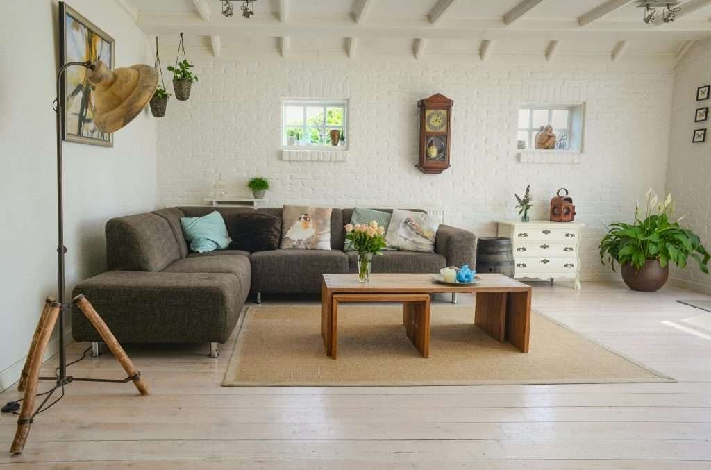 Bright and cozy living room with a brown sectional sofa, wooden coffee table, indoor plants, and white brick walls.
