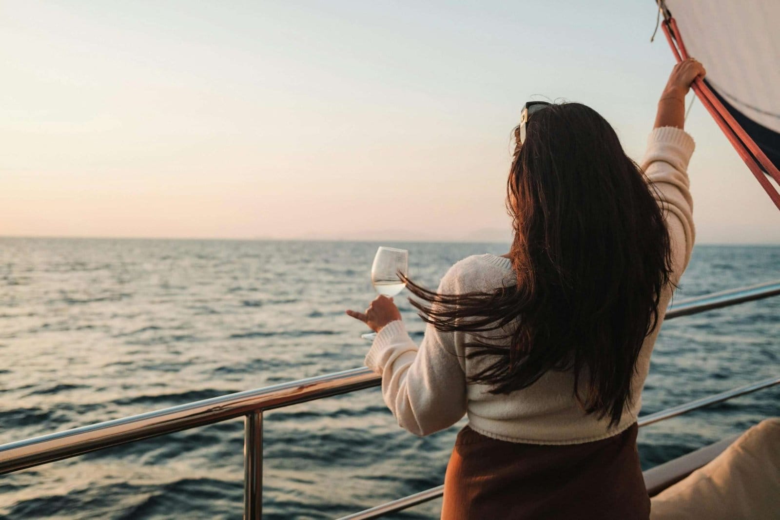 A woman standing on a cruise ship deck, holding a glass, gazing at the sunset over the ocean.